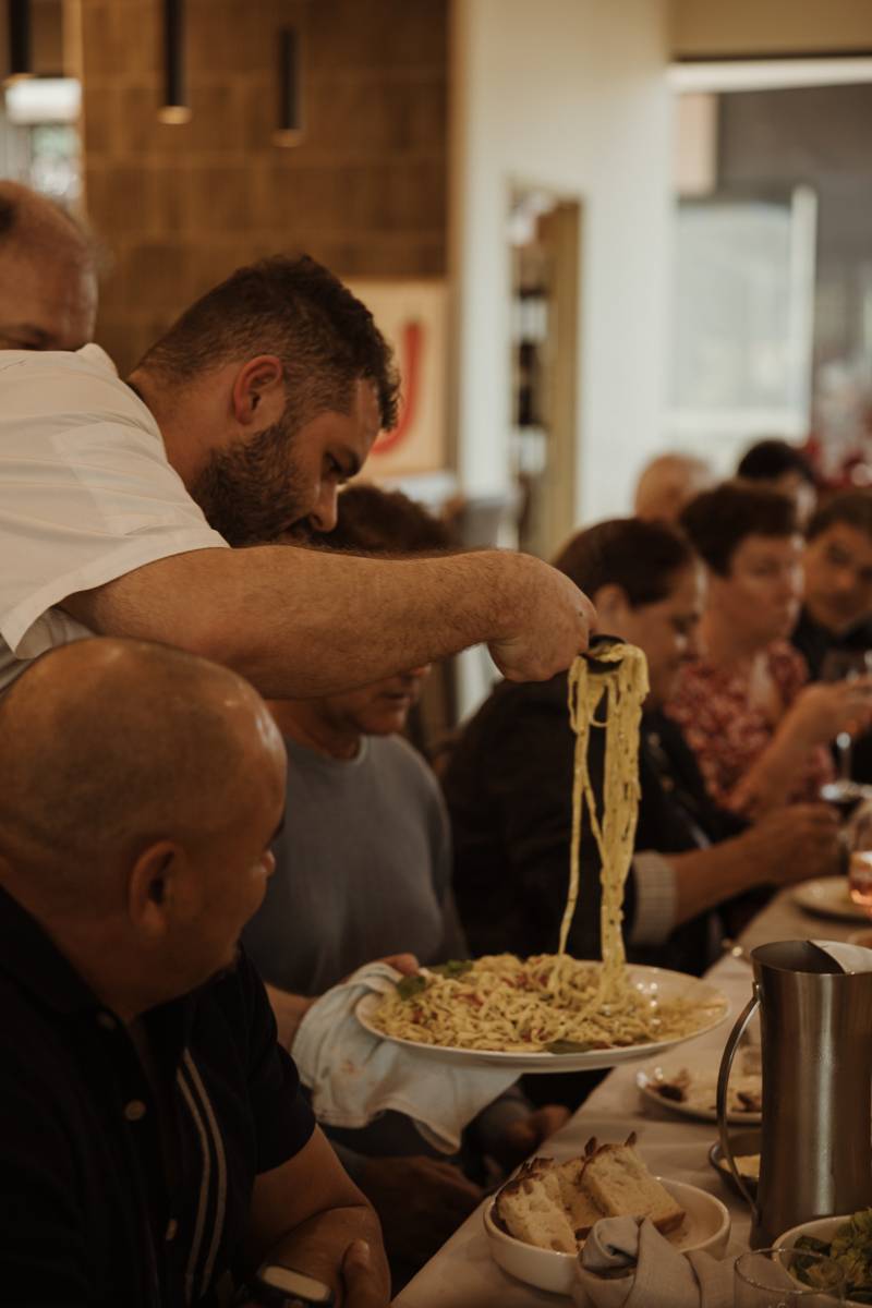 A chef lifting fresh pasta from a plate to serve guests at the table